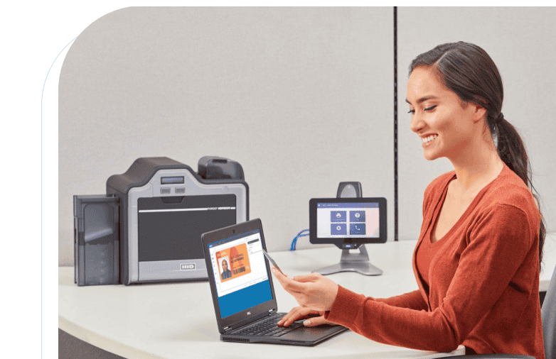 A woman sits at a desk holding an ID card, interacting with a laptop displaying ID information, with a card printer and monitor in the background.