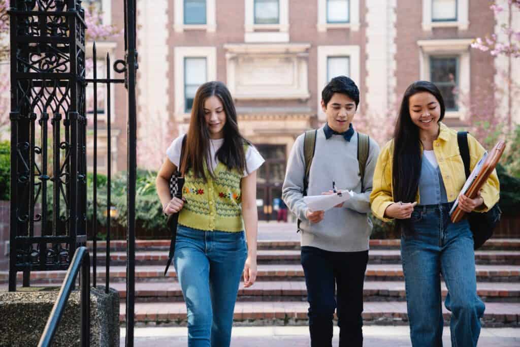Three students with backpacks walk together on a campus pathway, holding notebooks and papers, with a brick academic building in the background.