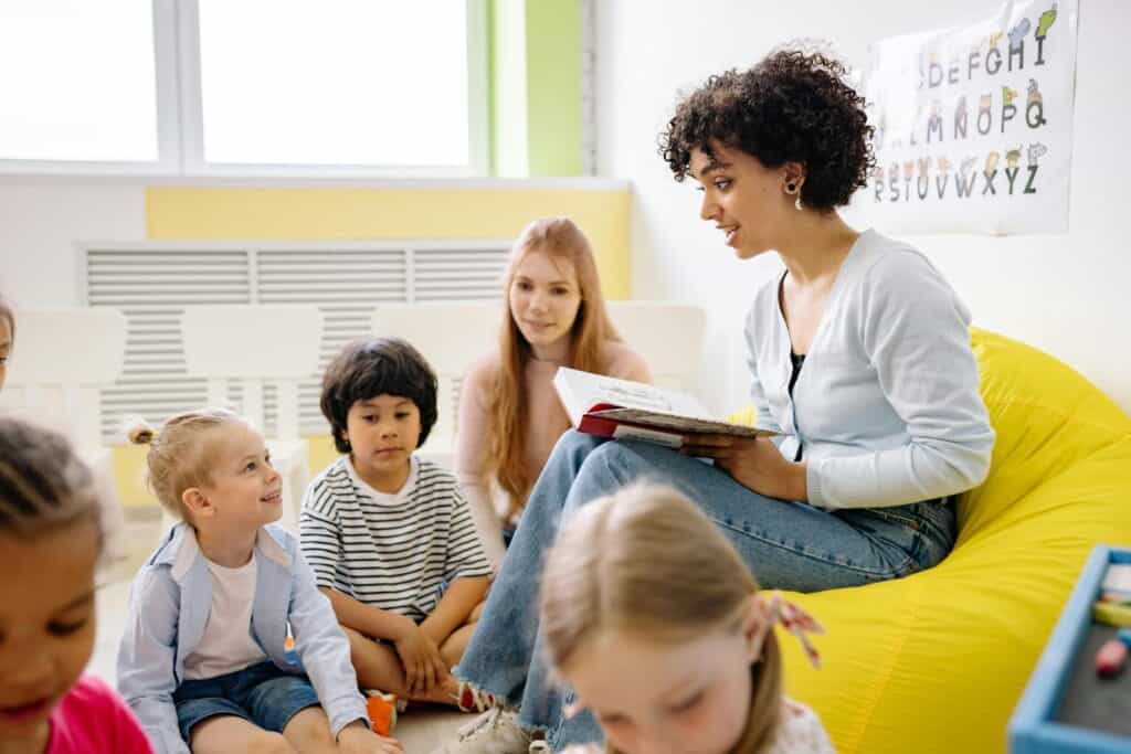 A woman reads a book to a group of young children sitting on the floor in a brightly lit classroom, while another adult observes.