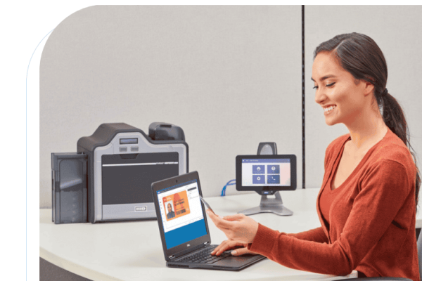 A woman sits at a desk holding an ID card, interacting with a laptop displaying ID information, with a card printer and monitor in the background.